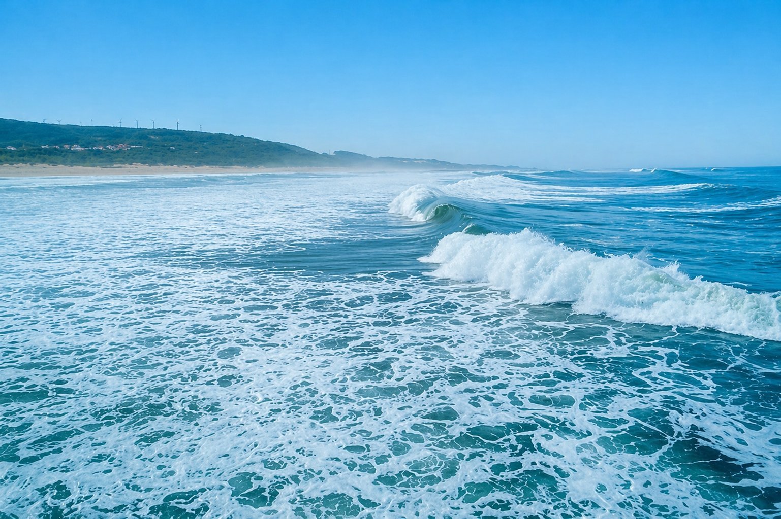 Ocean waves rolling onto a sandy beach under clear blue sky with hills visible in the distance