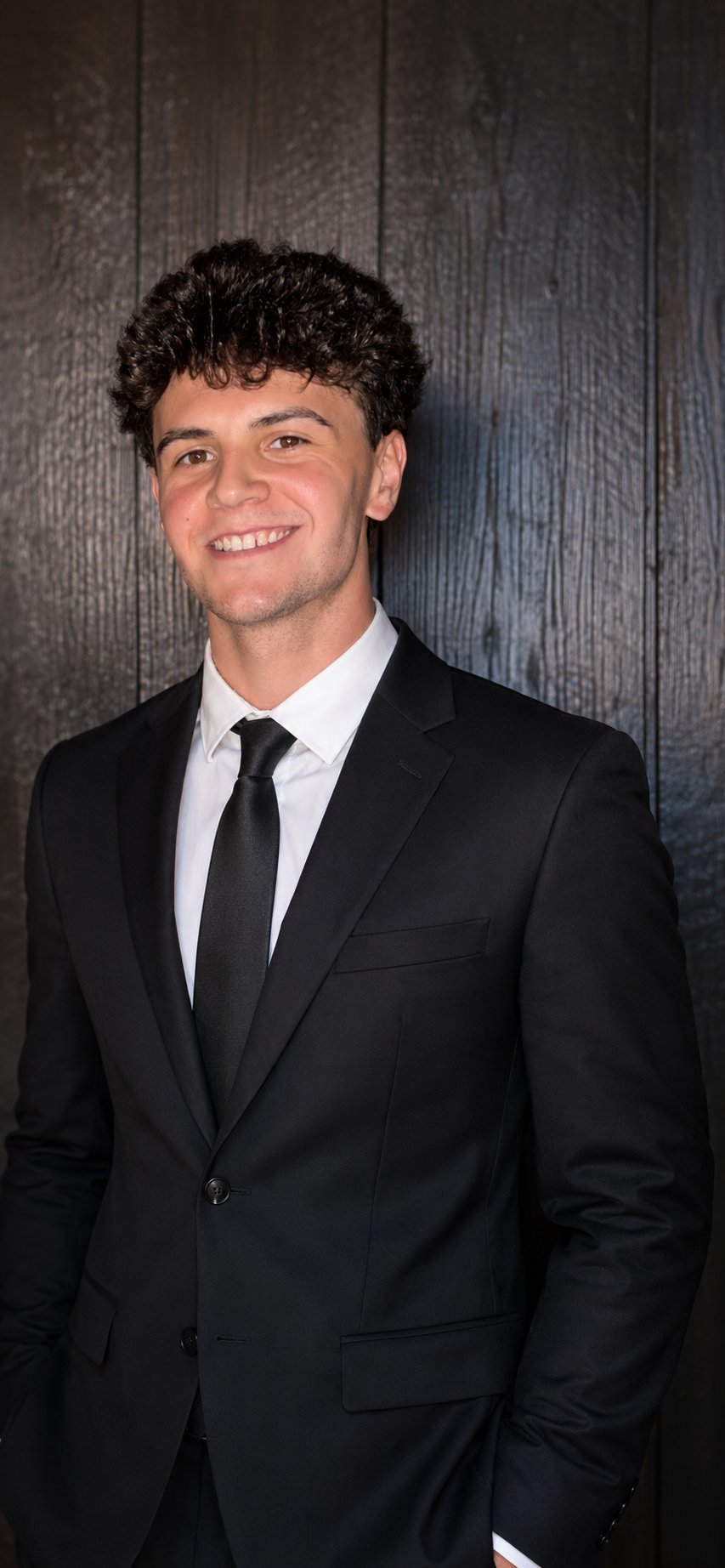 Young man in black suit and tie smiling at camera against dark wooden background